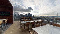 This expansive rooftop terrace offers a sophisticated urban retreat perched high above the city, delivering sweeping panoramic views of the surrounding skyline, waterfront, and iconic landmarks — including a prominent view of the Space Needle, suggesting a Seattle location. The space is thoughtfully divided into distinct outdoor and semi-covered zones designed for both relaxation and entertaining.

The open-air dining area features multiple sets of teak and metal tables and chairs arranged to maximize the breathtaking cityscape views, all framed by a sleek glass railing system. Adjacent to the dining zone, a generously sized covered lounge area is defined by striking warm-toned wood-paneled ceilings and large floor-to-ceiling sliding glass walls that blur the boundary between indoor and outdoor living. This sheltered section is furnished with substantial teak-framed sofas upholstered in deep navy blue cushions, complemented by glass-topped coffee tables and integrated overhead heating elements, ensuring year-round usability.

Structural columns clad in rich wood paneling add architectural character throughout, while large-format stone tile flooring unifies the entire terrace. Mounted televisions and built-in planters with ornamental grasses contribute to a polished, resort-inspired atmosphere. The overall ambiance is modern, refined, and inviting — an ideal venue for social gatherings with commanding views in every direction.
