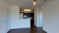 This single-floor space features an open-concept layout that seamlessly connects the living area to the kitchen. Dark hardwood floors run throughout, adding warmth and contrast against the crisp white walls. The kitchen is defined by dark espresso cabinetry, a black refrigerator, and built-in appliances, including a microwave and oven. A raised breakfast bar serves as a natural divider between the kitchen and the main living area, offering both functional counter space and a casual dining option. Overhead, a fluorescent light fixture illuminates the kitchen, while track lighting in the living area provides adjustable, directional light. A doorway with a paneled white door leads to an additional room, suggesting a private space such as a bedroom or bathroom beyond. HVAC vents are integrated into the ceiling, ensuring climate control throughout. The overall design aesthetic is contemporary and functional, with clean lines, neutral tones, and a straightforward layout that maximizes usable space.