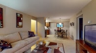 This single-floor residence offers a thoughtfully designed layout that connects living, dining, and kitchen areas in a cohesive, open flow. The living room features plush carpeting, a glass-top coffee table, and built-in wall shelving, creating a warm and functional gathering space. Adjacent to it, the dining area is anchored by a round glass table beneath a pendant light, with a framed mirror adding depth to the room. The galley-style kitchen is well-equipped, showcasing warm wood cabinetry, granite-style countertops, stainless steel appliances including a refrigerator and range, and hardwood-style flooring that adds a clean contrast to the rest of the carpeted unit. The bedroom is a tranquil retreat with soft carpeting, a window with drape treatments, and ample space for full bedroom furniture. A spacious walk-in closet with wire shelving and hanging rods provides generous storage. The bathroom features a granite-topped vanity with wood cabinetry and a large mirror. A dedicated utility room houses the washer and water heater, adding practical convenience. Neutral warm tones throughout create a cohesive and inviting ambiance.
