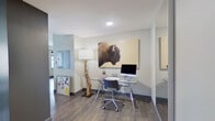 This thoughtfully designed one-bedroom, three-bathroom residence offers a refined blend of functionality and contemporary style across a single floor. The open-concept kitchen and living area serves as the heart of the home, featuring a sleek kitchen with gray shaker cabinetry, stainless steel appliances including a French door refrigerator, a tile backsplash, and a bold blue island with a white quartz countertop and bar seating. The adjacent living room is bright and inviting, with multiple large windows that flood the space with natural light and offer urban views. Hardwood-style flooring flows throughout the main areas, adding warmth and continuity. A dedicated office provides a productive workspace, complete with a glass-top desk, a sculptural driftwood floor lamp, and distinctive wall art. The primary bedroom offers plush carpeting, a ceiling fan, natural light from windows, and direct access to an en-suite bathroom and a spacious walk-in closet with built-in shelving. The bathrooms are elegantly finished with large-format tile flooring, a glass-enclosed walk-in shower with subway tile surround, dark wood vanities, and clean white countertops. The overall ambiance is modern and curated, with an urban sensibility throughout.