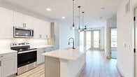 This well-designed one-bedroom, one-bathroom unit offers a thoughtful layout across a single floor, maximizing every square foot with clean lines and a contemporary aesthetic. Light wood-look flooring flows throughout the open-concept kitchen and living area, anchored by a large central kitchen island topped with white quartz and fitted with a matte black industrial-style faucet. The kitchen features crisp white shaker cabinetry, a marble-look tile backsplash, stainless steel appliances including a French door refrigerator and range, and a built-in microwave. A trio of geometric cage pendant lights adds a stylish focal point above the island. The living area extends naturally toward full-length French doors that open to a private balcony, allowing generous natural light to fill the space. A ceiling fan serves both the living and kitchen zones. The bedroom is carpeted for comfort and includes a ceiling fan and a large window with exterior views. It connects directly to the full bathroom, which is outfitted with a white quartz vanity, bold-framed mirror, matte black fixtures, and large-format tile flooring. A well-organized walk-in closet with built-in shelving and hanging rods is accessible through the bathroom. A dedicated entrance and closet complete this efficiently designed unit.
