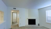 This single-floor space presents a straightforward and functional layout centered around a carpeted living area. The room is finished with clean white walls and a textured ceiling, giving it a bright, neutral appearance. A built-in fireplace with a simple white mantel serves as the focal point of the main living area, framed by a tiled hearth that adds a subtle contrast to the surrounding carpet. HVAC vents are positioned along the upper walls, ensuring climate control throughout the space. A hallway extends from the living area, lined with doors that lead to additional rooms, suggesting a practical and connected floor plan. The corridor is well-lit, with light filtering through from adjoining spaces, contributing to an open and accessible feel. The overall aesthetic is minimal and move-in ready, offering a neutral canvas that can be easily personalized. The combination of carpet in the living and hallway areas with tile at the fireplace hearth reflects a practical approach to flooring throughout the space.
