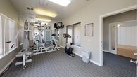 This fitness studio is designed to support a well-rounded workout routine in a comfortable, single-level space. The room features patterned carpet flooring throughout and is finished with neutral grey and white walls, giving it a clean, modern aesthetic. Overhead fluorescent lighting ensures the space is consistently well-lit, complemented by abundant natural light streaming through multiple large windows fitted with vertical blinds.

The cardio zone is equipped with a treadmill and two elliptical machines, positioned near the windows to allow exercisers to enjoy views of the surrounding outdoor environment. A multi-station strength training machine serves as the centerpiece of the space, offering a variety of resistance-based exercises from a single unit. A stationary cycle adds further variety to the available equipment.

A wall-mounted mirror expands the sense of space and allows users to monitor their form during workouts. A mounted television provides entertainment during exercise sessions. Storage is addressed through a metal wire shelving unit, and a waste bin is conveniently placed for general use. Posted fitness studio rules are displayed near the entrance, reinforcing a shared and respectful use of the facility. A frosted-glass door provides a discreet and accessible entrance to the studio, completing this practical and inviting fitness environment.