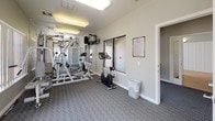 This fitness studio is designed to support a well-rounded workout routine in a comfortable, single-level space. The room features patterned carpet flooring throughout and is finished with neutral grey and white walls, giving it a clean, modern aesthetic. Overhead fluorescent lighting ensures the space is consistently well-lit, complemented by abundant natural light streaming through multiple large windows fitted with vertical blinds.

The cardio zone is equipped with a treadmill and two elliptical machines, positioned near the windows to allow exercisers to enjoy views of the surrounding outdoor environment. A multi-station strength training machine serves as the centerpiece of the space, offering a variety of resistance-based exercises from a single unit. A stationary cycle adds further variety to the available equipment.

A wall-mounted mirror expands the sense of space and allows users to monitor their form during workouts. A mounted television provides entertainment during exercise sessions. Storage is addressed through a metal wire shelving unit, and a waste bin is conveniently placed for general use. Posted fitness studio rules are displayed near the entrance, reinforcing a shared and respectful use of the facility. A frosted-glass door provides a discreet and accessible entrance to the studio, completing this practical and inviting fitness environment.
