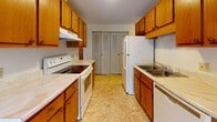 Located at 65 Carriageway Dr in Fitchburg, Massachusetts, this single-floor unit offers a practical and well-organized layout designed for comfortable everyday living. The open-concept living and dining area is generously sized, finished with neutral wall-to-wall carpeting and bathed in natural light from a large sliding glass door that opens to an outdoor balcony, as well as an additional window framing views of surrounding greenery. The galley-style kitchen is efficiently designed, featuring warm oak cabinetry that extends to the ceiling for ample storage, light-toned countertops, white appliances including an electric range and refrigerator, a stainless steel sink, and easy-care tile flooring. A pass-through peninsula connects the kitchen to the living area, enhancing the sense of openness. The bedrooms are carpeted and quiet, each offering built-in closets with louvered doors for organized storage. The full bathroom is finished with classic tile wainscoting, a combined tub and shower with a handheld showerhead, a vanity with an integrated sink, and small-format white floor tile. Baseboard heating is present throughout, and the overall palette of warm neutrals lends the space a clean, move-in-ready appearance.