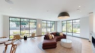 This contemporary single-floor communal lounge is defined by its bright, open design and an abundance of natural light streaming through expansive floor-to-ceiling windows with sleek black frames. The windows wrap generously around the main seating area, offering wide views of surrounding greenery and the outdoors, creating a seamless connection between the interior and exterior environments.

The primary lounge area centers around a curved sectional sofa in rich earth tones, complemented by a round stone coffee table and layered area rugs that add warmth to the light wood flooring. Swivel accent chairs in neutral upholstery are thoughtfully arranged to encourage conversation. A striking dome pendant light anchors the room visually from above.

Along one wall, a linear fireplace with a light brick surround and an integrated wood and stone wet bar provides a cozy focal point, adding both warmth and elegance to the space. Recessed lighting and subtle wall sconces contribute to a refined atmosphere throughout.

A secondary nook offers a more intimate setting, featuring a built-in banquette with velvet upholstery, open wood shelving with decorative accents, and a small café-style table with wooden chairs — ideal for casual meetings or quiet relaxation. Fluted wall panels and a slatted accent wall add refined textural detail to this area.

Overall, the space exudes a modern, welcoming ambiance suited for both socializing and unwinding.