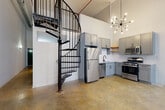 This industrial-chic loft masterfully blends raw architectural character with modern finishes. Soaring ceilings create an expansive, airy atmosphere throughout, while polished concrete floors provide a sleek, cohesive foundation across the living spaces. A striking black wrought-iron spiral staircase serves as a dramatic focal point, rising toward the upper level and adding a sculptural element to the open-plan layout.

The kitchen is a standout feature, outfitted with grey shaker-style cabinetry, granite countertops, a white subway tile backsplash, open wooden shelving, and a full suite of stainless steel appliances including a gas range, refrigerator, dishwasher, and microwave. A mid-century modern sputnik chandelier overhead adds a refined touch. An exposed brick wall with tall, arched windows floods the main living area with natural light, reinforcing the building's historic industrial origins.

Additional spaces include a dedicated laundry room with a full-size washer and dryer, a bathroom with a tub-shower combo, and utility areas with accessible electrical panel and climate control. The open living areas offer generous wall space, ideal for flexible furniture arrangements. Throughout, the interplay of original brick, crisp white walls, and industrial detailing creates a dynamic and inviting urban living environment.