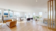 The Helios Lounge at 540 W Bay Terrace in Esquimalt, BC is a thoughtfully designed communal lounge that exemplifies contemporary west coast living. The single-floor space is defined by its crisp white palette, light hardwood flooring, and an abundance of natural light streaming through large, floor-to-ceiling windows that frame expansive views of the surrounding Esquimalt neighbourhood and beyond.

The generous open-plan living and dining area forms the heart of the lounge. The living zone is furnished with a plush sectional sofa, paired accent chairs, and a central coffee table arranged on a soft area rug — creating an inviting setting for both relaxed gatherings and social events. Warm-toned accent seating and curated artwork add character to the space, while a wall-mounted television provides a media focal point.

A sleek, minimalist kitchenette is tucked neatly to one side, featuring flat-front white cabinetry, a stainless steel sink, and built-in shelving — ideal for hosting. A warm wood slatted divider screen elegantly separates the dining area, which includes a light wood table and chairs set beneath views of the treetops.

A full bathroom serves the lounge, and glazed doors open directly onto a rooftop terrace, further extending the usable space and offering sweeping outdoor views. Recessed lighting throughout ensures the space remains bright and welcoming at any hour.