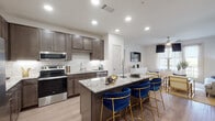 This single-floor residence is designed with an open-concept layout that seamlessly connects the kitchen, dining, and living areas. The kitchen is a standout feature, showcasing dark taupe shaker-style cabinetry, a classic white subway tile backsplash, stainless steel appliances, and a generous granite-topped island with seating for several bar-height chairs. Light wood-look flooring flows throughout the main living areas, enhancing the bright, airy atmosphere created by recessed lighting overhead.

The living room offers a welcoming retreat, anchored by a bold dark accent wall, access to an outdoor balcony, and natural light from large glass doors. The bedrooms feature plush carpeting, ceiling fans, and windows that bring in outdoor views. The primary suite connects to a well-sized bathroom with granite countertops, dark cabinetry, and a tub-shower combination finished with white tile. A dedicated walk-in closet with built-in shelving provides ample storage. A secondary bathroom mirrors similar quality finishes and also includes a walk-in closet. Overall, the residence balances contemporary design with practical, comfortable living spaces.
