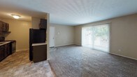 Located at 3375 Gibralter Heights Dr in Toledo, Ohio, this single-floor unit offers a straightforward and functional layout across several distinct areas. The living room is generously sized, featuring wall-to-wall carpeting in a neutral tone, warm beige walls, and large vertical-blind-covered windows that allow natural light to fill the space. Ceiling vents are positioned throughout, indicating a central HVAC system.

The kitchen is equipped with dark wood cabinetry, light countertops, and black appliances including a range, dishwasher, and refrigerator. A single-basin sink sits beneath the upper cabinets, and tile-style flooring runs through the kitchen and adjoining areas, providing a practical surface in high-traffic zones.

The bedroom area continues the carpeted flooring and neutral palette, with a window providing natural light, a closet, and access to the bathroom. The full bathroom includes a tub and shower combination with white tile surround, a toilet, and a vanity with a white cabinet, single-basin sink, and a lighted medicine cabinet mirror above.

The overall layout connects the living, kitchen, bedroom, and bathroom in a compact and efficient arrangement suited for everyday use.