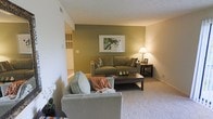 This single-floor residence centers around a well-appointed living room that combines comfort and style. The space is defined by a bold olive green accent wall, which serves as a focal point and adds depth to the room's neutral color palette of whites and beiges. A framed landscape artwork is displayed prominently on the accent wall, complementing the earthy tones throughout.

The living room is furnished with a taupe sofa adorned with patterned throw pillows, paired with a light grey armchair draped with a warm orange throw blanket, creating an inviting and layered seating arrangement. A dark wood rectangular coffee table anchors the seating area, styled with decorative accents. A matching side table holds a classic cream table lamp, providing warm ambient lighting that enhances the room's cozy atmosphere.

Wall-to-wall carpeting in a light neutral tone runs throughout the space, contributing to the comfortable feel underfoot. The white textured ceiling and light-toned walls allow the accent wall to stand out effectively. A large wall vent is visible, indicating a built-in HVAC system. The open layout suggests a natural flow between the living area and adjacent spaces, offering a functional and welcoming environment.
