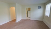 This single-floor unit at 216 B offers a clean, straightforward layout designed for comfortable everyday living. The main living area is finished with wall-to-wall carpet in a warm neutral tone, contributing to a cozy and quiet atmosphere. Soft green-tinted white walls keep the space feeling light and airy throughout.

A distinguishing architectural detail is the arched doorway on the left side of the living area, adding a subtle decorative touch to an otherwise understated design. White painted baseboards run along the perimeter of the room, providing a crisp contrast to the carpeted floors.

A standard rectangular doorway leads directly into the kitchen, which features tile flooring and wood-toned cabinetry. A white refrigerator is visible from the living area, indicating a functional kitchen setup. An electrical panel is also present on the wall of the main living area, offering practical accessibility.

Overall, the unit presents a modest and well-maintained living environment with a neutral palette that offers a versatile backdrop for personalization. The single-floor layout ensures ease of accessibility and straightforward navigation throughout the space.