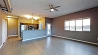 This single-floor space offers a thoughtfully designed open-concept layout that seamlessly connects the kitchen and living areas. The kitchen is a standout feature, outfitted with dark wood cabinetry, stainless steel appliances including a refrigerator and microwave, and a raised breakfast bar that serves as both a functional workspace and a natural divider between the kitchen and the main living area. Elegant pendant lights with amber glass shades hang above the bar, adding a warm, inviting glow that complements the space's modern aesthetic. The living area is generously sized, bathed in natural light streaming through large windows that offer views of the surrounding landscape. A ceiling fan with an integrated light fixture is centrally positioned overhead, providing both comfort and additional illumination. The flooring throughout is a rich, dark hardwood-style that adds depth and sophistication to the interior. The walls are finished in a contemporary two-tone palette of warm taupe and cool gray, creating a balanced and stylish ambiance. A sliding glass door leads to an outdoor balcony, further enhancing the connection between indoor and outdoor living. The overall atmosphere is clean, modern, and welcoming.