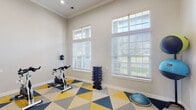 Located at 2801 E 120th Ave in Thornton, Colorado, this dedicated fitness and yoga studio offers a thoughtfully designed space for a wide range of exercise activities. The room is characterized by impressively tall ceilings accented with crown molding, creating an open and airy atmosphere that enhances the workout environment. Large windows with transom panels above flood the interior with abundant natural light, offering views of the surrounding greenery while keeping the space feeling fresh and energized. The flooring features a bold geometric carpet pattern in yellow, navy blue, and gray triangles, adding a vibrant, modern aesthetic to the room. Equipment includes multiple Spinning-brand cycling bikes, a full dumbbell rack, a rack of medicine balls, a BOSU balance trainer, resistance bands, and a wall-mounted flat-screen television for guided workouts or entertainment. A yoga mat station is also present, making the space versatile for stretching and mindfulness practices. Recessed lighting supplements the natural light, ensuring excellent visibility at any time of day. A frosted-glass French door provides access to an adjacent room, contributing to the overall polished and functional design of this community fitness amenity.