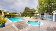 Located at 455 Crescent Ave in Sunnyvale, California, this outdoor recreational area offers a well-appointed pool facility designed for relaxation and leisure. At the heart of the space is a generously sized swimming pool with clear, blue-tinted water and tiled accents, providing ample room for both lap swimming and casual enjoyment. A dedicated pool house with sliding glass doors and a charming teal-accented entrance serves as a central amenity structure, offering storage and convenience for pool guests. Adjacent to the main pool sits a circular spa and hot tub, finished with decorative tiling and equipped with safety features including a mounted life ring. The surrounding deck area is furnished with rows of white lounge chairs and patio tables shaded by navy blue umbrellas, creating a comfortable outdoor retreat. Lush, mature trees frame the entire perimeter of the space, providing natural shade and a sense of seclusion from the surrounding neighborhood. Decorative potted plants, including striking tropical foliage, are placed throughout the patio to enhance the landscaping. A secured gate provides controlled access to the pool area, ensuring privacy and safety. The overall ambiance is that of a tranquil, resort-style outdoor amenity space well suited to the sunny California climate.