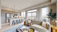 This single-floor residence offers a thoughtfully designed open-concept layout that seamlessly connects the main living spaces. The kitchen is a standout feature, showcasing flat-front cabinetry in a warm wood-tone finish, stainless steel appliances, and a generous island with a crisp white quartz countertop and bar seating. Pendant lighting and large windows flood the space with natural light, while light-toned hardwood flooring extends throughout, unifying the kitchen with the adjacent living area.

The living room is bright and welcoming, with oversized windows and access to an outdoor balcony offering open views of the surrounding area. Recessed lighting and neutral wall tones create a clean, modern atmosphere well-suited for both relaxation and entertaining.

The bedroom is a calm retreat, featuring plush carpeting, dual window exposure, and warm brass wall sconces flanking an upholstered headboard. A white dresser with a gold-framed mirror adds functional elegance to the room.

The bathroom is finished with dark marble-effect floor tiles, a wood-tone vanity with a white countertop, and a glass-enclosed walk-in shower detailed with elongated hexagonal tile. The overall aesthetic throughout the residence is contemporary and cohesive, emphasizing clean lines, neutral tones, and quality finishes.