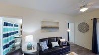 Located at 349 Campus Cove in Wilmington, North Carolina, this single-floor residence offers a cohesive coastal aesthetic that reflects the character of its surroundings. The living area serves as the heart of the home, featuring smooth white ceilings, clean wall lines, and warm hardwood-style flooring that grounds the space with a sense of natural comfort. A large, serene seascape painting anchors the main wall, complementing the nautical-themed décor and reinforcing the coastal ambiance throughout. The room is well-lit with a combination of natural light and ambient lighting, contributing to an open and airy atmosphere. Neutral wall tones provide a versatile backdrop that enhances the brightness of the space. A hallway visible from the living area suggests a thoughtful, connected layout that flows naturally between rooms. The overall design ethos leans into a relaxed, beach-inspired sensibility, with cohesive color palettes of navy, white, and soft earth tones. This single-level layout offers convenient, accessible living in a setting that balances comfort and style, making it well-suited for those seeking a calm and inviting coastal retreat in the Wilmington area.