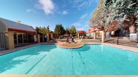 Located at 5350 Justin Dr NW in Albuquerque, New Mexico, this outdoor community pool area offers a well-appointed recreational retreat under the expansive New Mexico sky. The centerpiece is a generously sized swimming pool with clear blue water, complemented by a broad concrete sundeck that surrounds the pool on multiple sides, providing ample space for relaxation and recreation.

The deck is furnished with wicker-style lounge chairs and patio seating arrangements, including a table with a red umbrella, making it an ideal setting for outdoor gatherings. The pool area is enclosed by a sturdy black iron fence with masonry column accents, ensuring both security and safety. A life ring is mounted near the entrance gate, and posted signage notes that swimming is unsupervised.

A dedicated pool house structure with a metal roof and red exterior walls anchors one end of the space, providing a functional support facility. Mature trees are scattered throughout and beyond the fenced perimeter, offering natural shade and a lush backdrop that softens the built environment. The surrounding buildings feature a distinctive red adobe-inspired exterior that reflects the regional architectural character of Albuquerque. The entire space benefits from abundant natural sunlight and open sky views, creating a bright and inviting outdoor amenity.