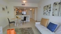 This well-appointed single-floor residence offers a cohesive open-plan layout that seamlessly connects the living, dining, and kitchen areas into one inviting space. The living area is furnished with a sleek white sofa accented by caramel leather cushions, complemented by a textured area rug and tropical wall art that adds a coastal character to the room.

The kitchen is modern and functional, featuring warm wood-toned cabinetry, a white refrigerator, and a suite of built-in appliances. A granite-topped breakfast bar with bar stools serves as a natural dividing element between the kitchen and the rest of the living space, offering both a casual dining option and additional prep area.

Adjacent to the kitchen, a compact dining area accommodates a glass-top table surrounded by contemporary transparent and dark accent chairs, ideal for everyday meals. The light-toned tile flooring runs consistently throughout, enhancing the sense of openness and continuity across all areas.

Recessed ceiling lighting provides even illumination throughout the space, while the clean white walls and ceilings contribute to the bright, airy ambiance. A hallway visible in the background leads to additional areas of the residence, suggesting a practical and well-organized floor plan overall.