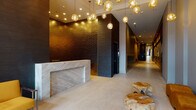 This elegantly designed lobby and corridor space makes a strong architectural statement through its carefully layered materials and refined aesthetic. The entry foyer draws immediate attention with its soaring double-height glazed entrance, flooding the space with natural light and providing views to the streetscape beyond. A substantial marble reception desk anchors the lobby, its veined stone surface contrasting beautifully against the dark, textured brick accent wall behind it. A striking cluster of amber globe pendant lights cascades from the ceiling, casting a warm, golden glow throughout the space and serving as a central design feature.

Natural organic elements are incorporated through raw, sculpted wood accent pieces used as decorative furniture, adding an earthy contrast to the polished finishes. A pair of mustard-yellow accent chairs provides a welcoming seating area near the entrance. Light-toned large-format tile flooring runs continuously from the lobby into an elongated corridor beyond.

The corridor is thoughtfully appointed with reclaimed wood wainscoting along the lower walls, built-in mailbox banks framed in warm wood, and a curated gallery of bold, colorful artwork displayed at intervals. Recessed lighting and accent spotlights illuminate the art while guiding visitors through the space, creating a gallery-like atmosphere that extends throughout.