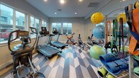 This single-level fitness center is thoughtfully designed to accommodate a full range of workout activities. The space is divided into two distinct functional zones: a cardio area and a strength training section, each outfitted with professional-grade equipment.

The cardio zone features treadmills, a spin bike, a rowing machine, and an elliptical trainer. Multiple windows along the exterior wall flood this area with natural light and provide views of the surrounding neighborhood, creating an energizing atmosphere. A large floor-to-ceiling mirror further amplifies the sense of openness.

The strength training area is anchored by a robust power rack system loaded with barbells and weight plates, complemented by a well-stocked dumbbell rack offering a wide range of weights. A bold typographic mural covers the back wall, displaying fitness-themed words such as "Strength," "Nutrition," "Physical," and "Athlete," reinforcing the motivational tone of the space.

Throughout the entire fitness center, a striking geometric-patterned floor in tones of gray, blue, and natural wood creates a visually dynamic foundation. Recessed ceiling lighting ensures consistent, even illumination across both zones, contributing to the clean, modern aesthetic of the overall space.