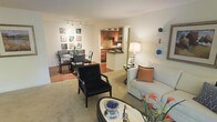 This single-floor residence offers a thoughtfully designed open-concept layout that seamlessly connects the living, dining, and kitchen areas into one cohesive, flowing space. The living area is comfortably proportioned, featuring neutral carpeting, a sofa with accent pillows, and a sleek oval coffee table that anchors the room. A bold accent armchair adds a striking contrast and visual interest to the seating arrangement. The dining area transitions naturally from the living space, centered around a round dining table with classic upholstered chairs and framed artwork adorning the walls nearby, complemented by a window that allows natural light to filter in. The kitchen is partially open to the main living area via a pass-through counter, showcasing rich warm-toned cabinetry, a tile backsplash, and a dark countertop breakfast bar. Track lighting overhead enhances the kitchen and dining zones, adding both functionality and ambiance. Decorative accents, including wall-mounted art pieces and colorful ceramic décor, lend a personalized, curated feel to the space. The overall ambiance is warm, inviting, and well-organized, making effective use of the available layout to create distinct yet connected living zones.