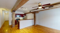 Located at 1329 Jefferson Street in Lynchburg, VA, this 1-bedroom, 1-bathroom unit showcases a compelling blend of historic industrial character and modern updates. Throughout the space, original heavy timber beams and structural columns are preserved and prominently displayed, lending a distinctive loft-like aesthetic that anchors the open layout.

The combined kitchen and living area forms the heart of the unit. The kitchen is outfitted with rich dark-wood cabinetry, granite countertops, stainless steel appliances — including a refrigerator, range, microwave, and dishwasher — and a peninsula with a warm wood-topped bar that separates the cooking space from the living area. Track lighting complements the kitchen's functionality.

The living area features warm-toned hardwood flooring, exposed brick accents along the lower walls, large windows that invite natural light, and a ceiling fan overhead. The bedroom continues the same hardwood flooring and timber beam details, with a ceiling fan and a double-door closet providing practical storage.

The bathroom is finished with tile flooring, a wood-toned vanity with an integrated sink, and a full tub-shower combination. A hallway connects the rooms, completing this well-appointed and characterful space.
