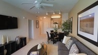 This single-floor residence showcases an open-concept design that integrates the living, dining, and kitchen areas into one cohesive, functional space. The living area is anchored by a plush, gray sectional sofa accented with decorative pillows, paired with a dark round coffee table — creating a comfortable and stylish gathering space. A ceiling fan with an integrated light fixture provides both airflow and ambient lighting overhead, while a track lighting system along the ceiling adds directed illumination across the space.

The dining area features a dark wood table surrounded by matching upholstered chairs, positioned adjacent to the kitchen for convenient access. Tall, lush indoor plants add a natural element and visual contrast against the warm beige walls. The kitchen is visible in the background, offering white cabinetry and appliances beneath additional overhead lighting.

Light-toned carpeting runs throughout the space, contributing to the warm and inviting ambiance. A hallway visible from the living area leads to additional rooms, and framed artwork along the walls adds personality. Overall, the space presents a clean, practical layout with tasteful furnishings and a cohesive neutral color palette.