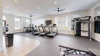 Located at 160 Bramble Ln in Lebanon, PA, this fitness center offers a well-appointed exercise space designed to meet a range of workout needs. The main exercise room is bright and spacious, benefiting from large windows that flood the area with natural light and offer views of the surrounding landscape. Dual ceiling fans and recessed lighting provide comfortable, well-regulated conditions throughout. The rubber-floored workout area is equipped with multiple treadmills, elliptical machines, a rowing machine, and a cable strength system, along with a full dumbbell rack, weight benches, stability balls, and wall-mounted mirrors that enhance the sense of openness. A wall-mounted television adds convenience for cardio sessions. The facility includes two bathrooms, each finished with large-format neutral ceramic tile on both the walls and floors. Both restrooms are designed with accessibility in mind, featuring black matte grab bars, open roll-in shower areas with floor drains, and hands-free fixtures. A water fountain and hand sanitizer station are conveniently located near the restroom entrance. The overall design is clean and modern, with a crisp white and blue color palette that contributes to a fresh, motivating atmosphere throughout the facility.