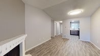 This single-floor unit is designed with a practical, open layout that connects the main living spaces efficiently. The living room serves as a central gathering area, highlighted by a corner fireplace with a white surround and a large sliding glass door that fills the space with natural light and provides access to an outdoor area. The flooring throughout the living and dining areas consists of light-toned, wood-style vinyl planks, complementing the neutral gray walls and textured ceilings.

The adjacent dining area flows naturally toward the kitchen, which features dark taupe cabinetry, light countertops, a black electric range with overhead ventilation, and a window above the sink. The layout provides functional counter and storage space for everyday use.

The bedrooms are carpeted in a neutral gray tone and each include windows with exterior views, offering natural light and a calm atmosphere. The bathroom is well-appointed with a dark wood vanity, a white countertop with an undermount sink, a medicine cabinet mirror, and a distinctive hexagon-patterned tile floor. A four-bulb vanity light fixture provides ample illumination.

Overall, the unit presents a clean, updated aesthetic with consistent neutral tones throughout.
