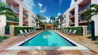 Located at 16185 Northwest 64th Avenue in Miami Lakes, Florida, this outdoor amenity space offers a beautifully maintained resort-style pool area that embodies the relaxed, tropical character of South Florida living. The centerpiece is a large, rectangular swimming pool with crystal-clear blue water, framed by an expansive terracotta-tiled sun deck that provides ample room for lounging and outdoor activities. Rows of cushioned chaise lounge chairs line both sides of the pool, offering residents and guests comfortable spaces to unwind and soak in the Florida sunshine. At the far end of the pool, a charming covered gazebo with a distinctive red-tiled roof serves as a shaded retreat, adding a Mediterranean architectural touch to the overall aesthetic. The space is enveloped by tall, swaying palm trees and dense tropical greenery, creating a lush, private atmosphere that enhances the resort-like ambiance. The surrounding multi-story buildings, featuring balconies overlooking the pool area, frame the courtyard and contribute to the sense of a well-integrated, community-centered outdoor environment. Classic lantern-style light posts are positioned throughout the deck, ensuring the space is welcoming both day and evening. The combination of architectural detail, thoughtful landscaping, and generous open-air layout makes this pool area a standout communal feature.