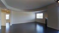 This single-floor space showcases a thoughtfully designed open living area with several distinctive architectural details. The most notable feature is the curved tray ceiling, which adds a soft, elegant dimension to the room and contributes to a sense of openness and flow. The smooth, neutral-toned walls complement the ceiling design, creating a cohesive and understated aesthetic throughout.

Underfoot, dark hardwood-style flooring anchors the space with a rich, polished finish that contrasts beautifully with the light walls and ceiling. A window fitted with horizontal blinds allows natural light to filter into the room, brightening the interior while offering privacy control. To one side, a fireplace is partially visible, suggesting a cozy focal point well-suited for a living or lounge area.

The overall layout is open and unfurnished, allowing the architectural elements to speak for themselves. The clean lines, curved ceiling detail, and dark flooring combine to give the space a modern yet inviting character. The single-floor layout ensures easy accessibility and a straightforward flow throughout the unit.