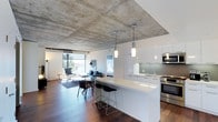 This contemporary single-floor space showcases a striking open-concept layout that seamlessly blends the kitchen, dining, and living areas into one cohesive living environment. The defining architectural feature is the expansive exposed concrete ceiling, which lends an industrial yet refined character to the space. Rich, dark hardwood flooring runs throughout, providing a warm contrast to the cool tones of the concrete overhead.

The kitchen is sleek and modern, outfitted with crisp white cabinetry, a clean-lined white quartz countertop, and stainless steel appliances including a built-in microwave and range. A peninsula with bar seating serves as both a functional workspace and a casual dining spot, complete with contemporary black bar stools. Pendant lighting in minimalist cylindrical forms hangs above, providing focused illumination.

The open living area beyond the kitchen benefits from floor-to-ceiling windows that flood the space with natural light and offer views of the surrounding landscape. The living zone is furnished with a comfortable sofa, accent chairs, and a dining table, creating distinct yet connected areas for relaxation and gathering. Track lighting along the ceiling complements the pendant fixtures, ensuring the space is well-lit throughout. Overall, the design balances industrial elements with modern sophistication.
