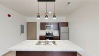 This thoughtfully designed 2-bedroom, 1-bathroom residence combines modern aesthetics with functional living across a single, efficiently organized floor. The open-concept kitchen serves as a central feature, showcasing rich dark-walnut cabinetry, marble-look countertops, and a large island with a double-basin sink. A trio of stylish pendant lights with gold-accented fixtures illuminates the island, while a full suite of stainless steel appliances — including a refrigerator, electric range, and over-the-range microwave — completes the kitchen. Light wood-look flooring flows throughout the unit, lending warmth and continuity to every room. The open living area benefits from natural light through large windows that frame outdoor views. Both bedrooms are well-proportioned, with the primary bedroom offering generous square footage, and each room featuring clean white walls and wood-trimmed accents. The primary bedroom includes a closet with sliding doors providing practical storage. The bathroom features dark cabinetry with marble-look counters, a single vanity sink, and a tall linen storage tower. The overall design is clean and contemporary, creating a bright, move-in-ready living environment.