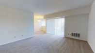 Located at 2000 Maplewood Dr in York, Pennsylvania, this single-floor residence offers a comfortable and straightforward layout designed for easy everyday living. The living area is generously sized, finished with wall-to-wall neutral carpet that lends warmth underfoot and creates a cohesive, inviting atmosphere throughout. Large vertical-blind-covered sliding glass doors dominate one wall of the living space, allowing an abundance of natural light to filter in while providing direct access to the outdoors. A built-in wall vent ensures year-round climate comfort. The living area flows seamlessly into an adjacent dining space, which is distinguished by its own set of vertical blinds and a classic chandelier overhead, adding a touch of elegance to the transition between spaces. Clean white walls and light-toned ceilings keep the interior feeling bright and airy. The open connection between the living and dining areas encourages a natural flow, making the layout well-suited for both daily routines and casual entertaining. The overall ambiance is clean, neutral, and move-in ready, offering a versatile canvas for a variety of interior styles and furnishings.