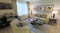 This single-floor space offers a thoughtfully designed living area that balances comfort and contemporary style. The living room serves as the centerpiece of the space, bathed in natural light streaming through a large window dressed with vertical blinds and flowing gold curtains, creating a warm and inviting atmosphere. The room is furnished with a cream-toned sofa adorned with decorative blue and grey patterned pillows, complemented by a matching woven accent chair featuring a floral cushion. A sleek glass-topped coffee table with a distinctive curved metal frame anchors the seating arrangement, which is set atop a textured area rug. A tall floor lamp provides additional ambient lighting, while a trio of framed botanical artwork arranged neatly on the wall adds a refined, curated touch to the neutral-toned walls. A under-window HVAC unit blends unobtrusively into the layout, ensuring year-round comfort. The overall ambiance of the space is polished and serene, with a cohesive color palette of creams, blues, and greens that lends a calm, sophisticated character throughout.