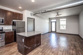 This single-floor unit showcases an industrial-modern aesthetic throughout its open layout. The kitchen serves as the centerpiece of the main living area, featuring dark espresso cabinetry, a full suite of stainless steel appliances including a refrigerator, range, microwave, and dishwasher, complemented by grey countertops and an island with additional storage. Open floating shelves add a contemporary touch alongside the cabinetry. A sliding barn door provides access to an adjoining space, adding a stylish functional element to the design.

Exposed cylindrical ductwork runs prominently across the ceiling of the main living area, reinforcing the industrial character of the space. Rich, dark hardwood-style flooring flows consistently throughout, providing visual continuity. The living area opens toward a door leading to an outdoor area, with windows allowing natural light to filter in, and a ceiling fan overhead.

The bedrooms are generously proportioned, each featuring ceiling fans, large windows with views to the outdoors, and access to bathrooms equipped with vanities and walk-in closets with built-in wire shelving systems. Dark interior doors with modern hardware maintain a cohesive, contemporary design palette throughout the unit.