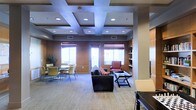 This single-floor communal space is designed to accommodate a variety of activities and social interactions through thoughtfully arranged seating zones. The room is anchored by a striking coffered ceiling with recessed lighting, which adds architectural depth and a sense of elegance to the space. Warm wood paneling along the walls creates a welcoming, refined ambiance throughout.

Natural light flows generously into the room through a series of large windows fitted with adjustable shades, brightening the neutral carpeted flooring and contributing to an open, airy feel. A central lounge area features a dark leather sofa paired with a glass coffee table, offering a comfortable gathering spot. Adjacent to this is a casual reading or accent seating area with a classic upholstered armchair. Toward one side of the room, a dining or collaboration zone is outfitted with round tables and lightweight chairs, ideal for meetings or informal gatherings.

A built-in bookcase provides both functional storage and a decorative element along one wall, while structural columns are seamlessly integrated into the design. The overall layout balances comfort and functionality, making this space well-suited for both quiet, individual use and larger group interactions.