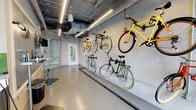 This single-level bike storage and maintenance room is purpose-built to accommodate cyclists with both practicality and organization in mind. The space features a series of wall-mounted bike racks lining one side, allowing multiple bicycles to be stored efficiently — both at floor level and elevated positions — maximizing the use of vertical space. The concrete walls and polished concrete flooring give the room a clean, utilitarian aesthetic that is easy to maintain.

Overhead, exposed ductwork and conduit run along the ceiling alongside sleek linear LED light fixtures, providing bright, even illumination throughout the space. A dedicated bike repair and maintenance station is positioned near the entrance, equipped with tools and a floor-standing bike stand, making it convenient for residents or users to perform routine upkeep on their bikes.

The room is accessed through a standard door, with what appears to be a glass-partitioned area near the entry point, possibly serving as a secure access vestibule. The overall ambiance is functional and well-organized, reflecting a thoughtfully designed amenity space built to serve an active, bike-friendly community.