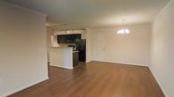 This single-floor residence features an open-concept layout that seamlessly connects the kitchen and living areas, creating a cohesive and functional living environment. The kitchen is appointed with dark espresso-toned cabinetry that contrasts sharply against the neutral, light-colored walls, lending the space a bold and modern aesthetic. Black appliances, including a refrigerator, range, and microwave, complement the cabinetry, while a peninsula with a raised bar provides both additional workspace and a natural visual divider between the kitchen and the rest of the open floor plan. A track lighting fixture over the kitchen ensures practical illumination for meal preparation. The living area is generously proportioned, offering ample room for a variety of furniture arrangements. Rich, warm-toned hardwood-style flooring runs throughout, anchoring both spaces with a consistent and inviting feel. An elegant chandelier with candelabra-style bulbs hangs over the living area, adding a refined decorative touch. Crown molding detail along the upper walls contributes an additional layer of architectural character. The overall ambiance is clean, open, and move-in ready.