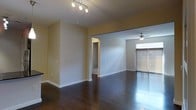This single-floor space presents an open, flowing layout anchored by rich, dark hardwood flooring that runs throughout the main living areas. The interior is organized around a spacious main living zone that transitions seamlessly into a secondary room through an open archway, creating a connected yet defined floor plan. The main area features a modern track lighting fixture mounted to the ceiling, providing adjustable, directional illumination across the space. The adjoining room is equipped with a ceiling fan, adding both functionality and a casual architectural element. A large sliding glass door with shutters allows natural light to pour into the secondary area, brightening the interior and offering access to the outdoors. Neutral, warm-toned walls complement the dark flooring, creating a balanced contrast throughout. White baseboards provide clean visual definition along the perimeter of each room. A hallway visible through an interior doorway suggests additional rooms beyond the main living space. The overall ambiance is clean, understated, and versatile, lending itself well to a variety of furniture arrangements and interior design styles.