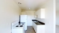 This single-floor space is designed with practicality and functionality at its core. The kitchen serves as a central feature, offering a clean and efficient layout that maximizes the available space. White cabinetry lines the walls, providing ample storage through both upper and lower cabinets, complemented by an open shelf for easy-access items. The dark countertops offer a subtle contrast to the bright white surfaces, creating a cohesive and tidy aesthetic. A double-basin sink is conveniently positioned along the counter, ideal for everyday use. The kitchen is equipped with a full suite of white appliances, including a refrigerator and a coil-burner electric range, ensuring all essential cooking and food storage needs are met. The neutral-toned tile flooring is both durable and easy to maintain, well-suited for a high-use area. The light-colored walls and ceiling, paired with a ceiling vent and overhead lighting, contribute to a bright and airy atmosphere throughout the space. The overall design is straightforward and move-in ready, making it a practical choice for those seeking a no-fuss, well-equipped living environment.