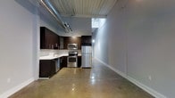 This single-floor urban loft-style space combines industrial design with modern finishes to create a sleek, contemporary environment. The open-concept layout is anchored by polished concrete floors that reflect light and lend an understated sophistication throughout. Exposed silver ductwork runs along the ceiling, complementing the raw, industrial aesthetic while adding to the sense of vertical space and openness above.

The kitchen is a standout feature, outfitted with rich dark cabinetry that contrasts sharply against light countertops and a clean white backsplash. A full suite of stainless steel appliances — including a refrigerator, electric range, built-in microwave, and dishwasher — provides a fully functional and stylish culinary space. Track lighting overhead ensures excellent task illumination throughout the kitchen area.

Adjacent to the kitchen, the open living area offers generous room for flexible use, bathed in natural light that filters in through a tall vertical window, creating a bright and airy atmosphere. The smooth grey walls and clean white baseboards maintain a neutral palette that allows the architectural details to take center stage. Overall, this space delivers a cohesive industrial-modern aesthetic, ideal for those who appreciate open, adaptable, and design-forward living environments.