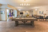 Located at 590 Downey Green St in Hampton, Virginia, this single-floor community clubhouse is designed to foster social interaction and relaxation in a stylish, welcoming environment. The space is anchored by wide-plank hardwood-style flooring that flows seamlessly throughout, tying together the various distinct zones within the open layout.

Upon entry, a refined lounge area greets visitors with contemporary furnishings, including plush sofas, accent chairs, and a striking sputnik chandelier overhead, all set against clean white walls adorned with colorful abstract artwork. Recessed lighting and natural light from large windows and glass-panel doors contribute to the bright, airy atmosphere.

The central gathering area expands into a generously proportioned open space featuring multiple conversation nooks, a wall-mounted fireplace display, and a large flat-screen television — ideal for both casual lounging and group entertainment. A full-size pool table anchors the game area, adding a lively recreational focal point.

A well-appointed kitchenette sits along one wall, outfitted with rich dark cabinetry, a tiled backsplash, ample counter space, a dishwasher, and a full refrigerator, alongside a dining area with seating for gatherings. French doors provide additional access points, connecting the interior to outdoor areas and allowing natural light to flood the space throughout the day.