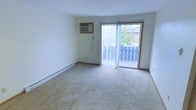 This single-floor space features a well-proportioned living area finished with neutral wall-to-wall carpeting that adds warmth underfoot. The room is kept bright and open by a full sliding glass door fitted with vertical blinds, which provides direct access to a private outdoor balcony enclosed by a white railing. The balcony overlooks a lush green tree canopy, offering a pleasant natural view and allowing ample natural light to filter into the interior. Climate control is handled by a wall-mounted air conditioning unit positioned above baseboard heating that runs along the perimeter of the room, ensuring year-round comfort. The walls are finished in a clean, neutral white tone, providing a versatile backdrop suitable for a variety of interior styles. The overall layout is straightforward and functional, with an uncluttered floor plan that maximizes usable living space. The combination of natural light from the sliding door, the outdoor connection via the balcony, and the practical climate control features makes this a comfortable and adaptable living space.
