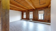 Located at 24 Park St in downtown Springfield, MA, this 2-bedroom, 1-bathroom loft-style unit showcases a striking blend of historic industrial character and modern finishes. The spacious living area is defined by soaring wood-plank ceilings with exposed timber beams, original exposed brick walls, and light hardwood-style flooring. Arched brick windows allow generous natural light to pour in, offering views of the surrounding urban streetscape. Sturdy wooden support columns add to the authentic industrial aesthetic of the open layout. The kitchen is accessed through a graceful arched entryway and features warm wood cabinetry, dark granite-style countertops, and black appliances including a refrigerator, dishwasher, and range. Two kitchen areas provide ample prep and storage space. The bedrooms continue the loft aesthetic with exposed brick accent walls, wood beam ceilings, and soft carpeting underfoot. The primary bedroom is generously sized and includes multiple closets for ample storage. Additional closets are found throughout the unit. The full bathroom includes a clean, white tub-shower combination. With four closets total across the unit, storage is plentiful. This character-rich space offers a unique urban living experience rooted in architectural history.