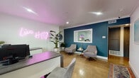 Located at 13751 Edwards St in Westminster, CA, this single-floor professional suite combines modern design with a warm, inviting atmosphere. The reception area immediately sets the tone with a glowing pink neon "Live Happy" sign, a bold teal accent wall, polished concrete flooring, and a contemporary front desk with a vibrant purple surface. Comfortable gray lounge chairs and lush tropical plants create a welcoming environment for clients.

Adjacent to reception, a windowed interior pass-through connects to a private consultation or therapy room, which features the same deep teal walls, an upholstered accent chair, a globe pendant floor lamp, and a round side table — offering an intimate, calming setting. Tasteful framed artwork and a patterned runner rug add character to the hallway transition between spaces.

The private office is well-appointed with an L-shaped wood-toned desk, computer workstation, built-in storage cabinetry, and natural light streaming through slatted blinds. A side door provides convenient secondary access.

The compact kitchen is fully equipped with gray shaker cabinetry, granite countertops, black appliances including a gas range and dishwasher, and a window overlooking the exterior. A small dining area with a bright teal chair completes the space. Crown molding and recessed lighting are consistent throughout, lending a cohesive, polished finish to the entire suite.