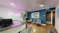 Located at 13751 Edwards St in Westminster, CA, this single-floor professional suite combines modern design with a warm, inviting atmosphere. The reception area immediately sets the tone with a glowing pink neon "Live Happy" sign, a bold teal accent wall, polished concrete flooring, and a contemporary front desk with a vibrant purple surface. Comfortable gray lounge chairs and lush tropical plants create a welcoming environment for clients.

Adjacent to reception, a windowed interior pass-through connects to a private consultation or therapy room, which features the same deep teal walls, an upholstered accent chair, a globe pendant floor lamp, and a round side table — offering an intimate, calming setting. Tasteful framed artwork and a patterned runner rug add character to the hallway transition between spaces.

The private office is well-appointed with an L-shaped wood-toned desk, computer workstation, built-in storage cabinetry, and natural light streaming through slatted blinds. A side door provides convenient secondary access.

The compact kitchen is fully equipped with gray shaker cabinetry, granite countertops, black appliances including a gas range and dishwasher, and a window overlooking the exterior. A small dining area with a bright teal chair completes the space. Crown molding and recessed lighting are consistent throughout, lending a cohesive, polished finish to the entire suite.