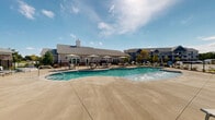Located at 458 E Riviera Blvd in Foley, Alabama, this outdoor amenity space centers around a generously sized freeform swimming pool designed to evoke a resort-style atmosphere. The pool features a zero-entry wading section with gradual steps and blue tile accents, making it accessible and inviting for all ages. An ADA-compliant pool lift is also present, ensuring full accessibility. A separate shallow splash zone is connected to the main pool, ideal for younger guests.

Surrounding the pool is an expansive concrete sun deck furnished with lounge chairs and chaise seating, perfect for relaxing in the Alabama sunshine. Multiple umbrella-shaded tables and chairs provide comfortable outdoor dining and gathering areas adjacent to the pool. A single-story clubhouse building with a pitched roof, cupola accent, and wall-mounted lantern lighting anchors one side of the deck, offering covered access and interior amenities nearby.

The entire pool area is enclosed by a black iron fence, providing both safety and an open sightline to the surrounding multi-story residential buildings finished in blue and gray siding. A playground structure is also visible within the community grounds. The overall ambiance is bright, open, and community-focused, designed to serve as a central outdoor gathering hub under the wide open skies of coastal Alabama.