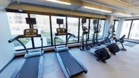 This fitness room offers a practical and well-organized space designed for cardio-focused workouts. The room is lined with large, multi-pane windows that span nearly the full length of one wall, flooding the interior with natural light and offering open views of the surrounding outdoor environment, including greenery and adjacent buildings. The bright, airy atmosphere is further enhanced by overhead fluorescent lighting, ensuring consistent illumination throughout the space. The flooring consists of durable, cushioned tiles in a neutral tone, well-suited for high-traffic fitness use. A row of treadmills is positioned directly in front of the windows, allowing users to enjoy the outdoor scenery during their workouts. Alongside the treadmills, multiple elliptical cross-trainers are arranged to maximize the use of available floor space. Each cardio machine is equipped with an individual monitor, supporting entertainment and workout tracking. The exposed ceiling structure adds an industrial aesthetic element to the otherwise clean and functional design. Overall, the space presents a focused and motivating environment suited for regular cardiovascular exercise.